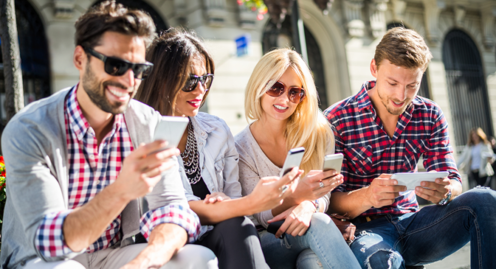 A group of young adults sitting outdoors, smiling and deeply engaged with their phones and tablets, representing users who evolve from casual adoption to confident, high-value power usage.